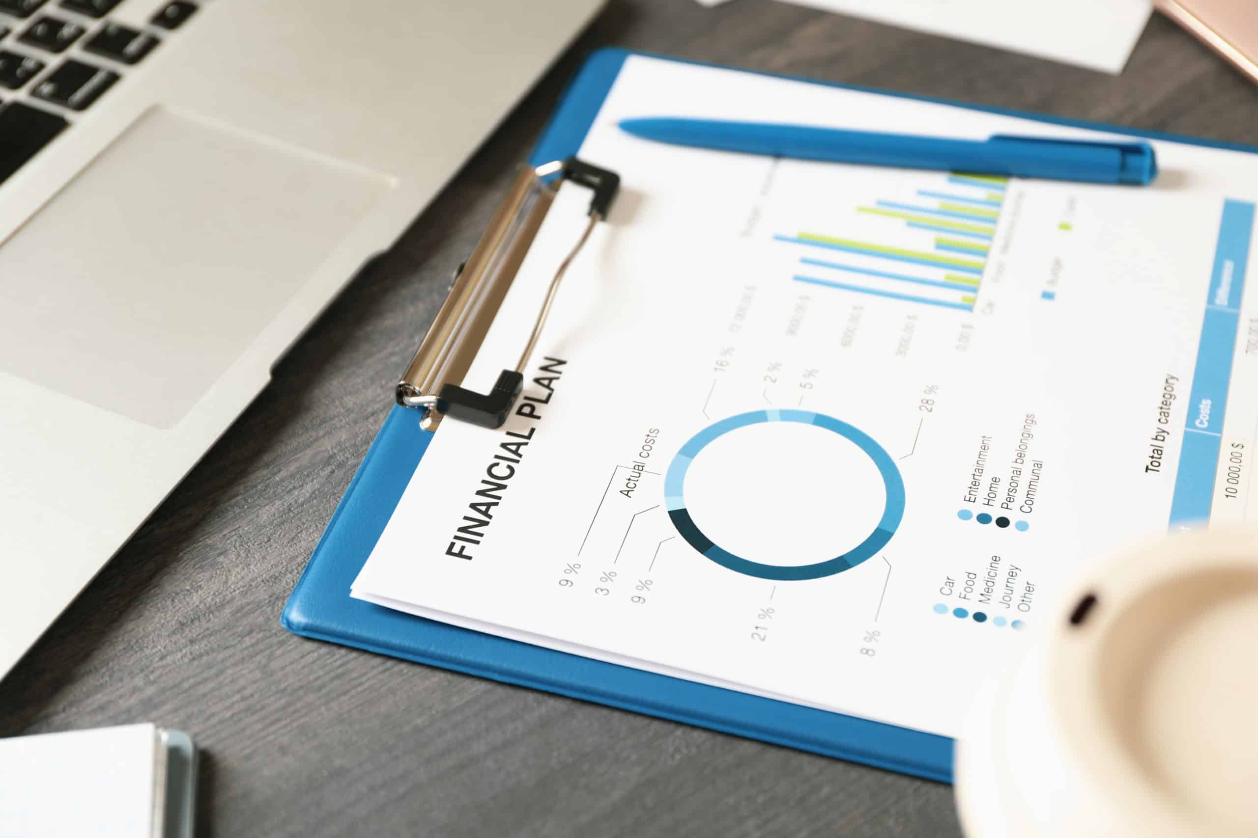 Financial plan with charts on clipboard next to laptop, pen, and coffee cup on a desk.