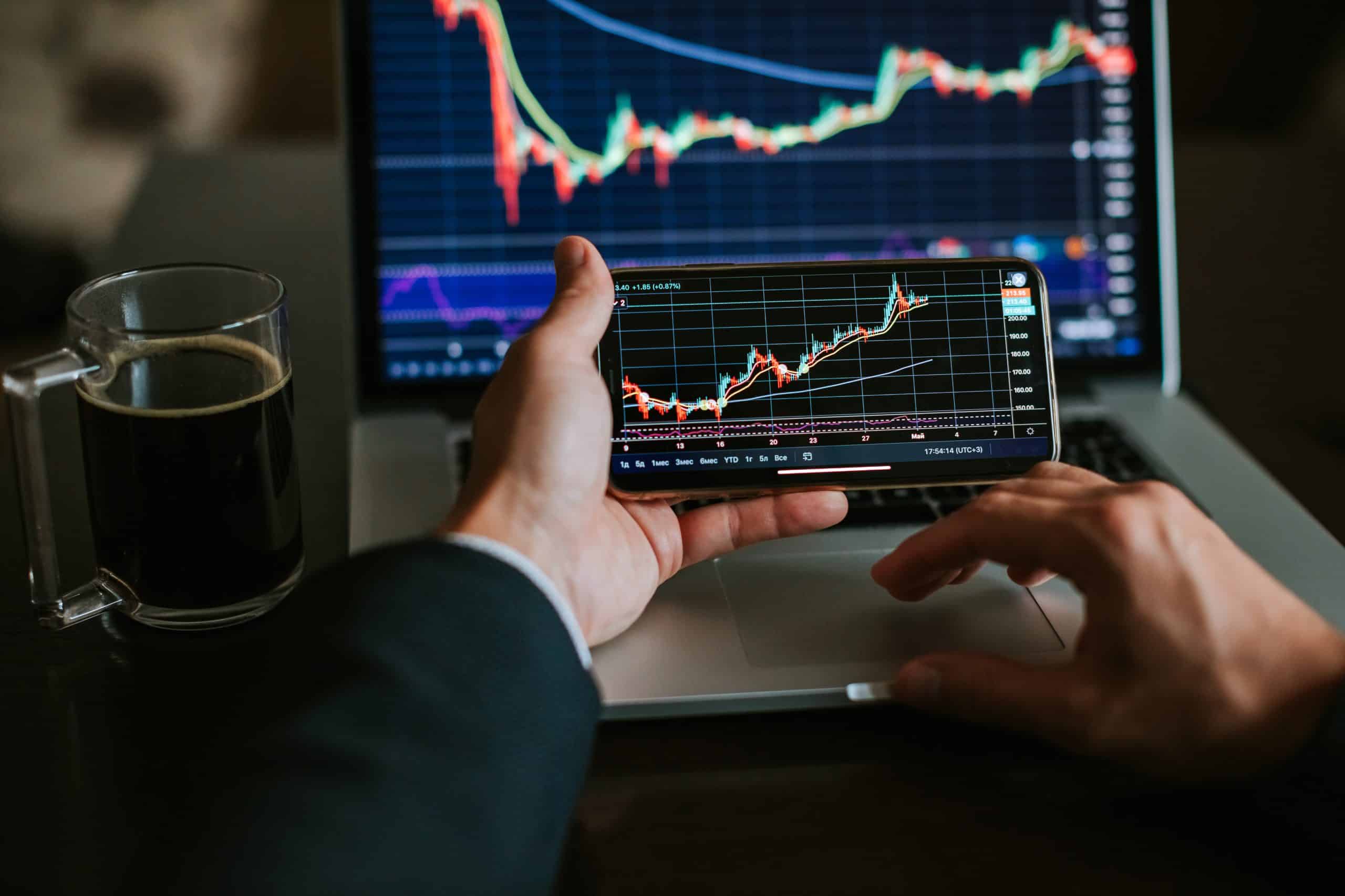 Person analyzing stock charts on phone and laptop with coffee mug.