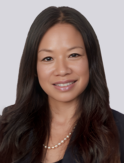 Smiling woman with long dark hair wearing a suit jacket and necklace, in front of a light gray background.