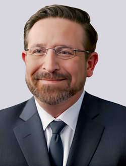 Smiling man in a business suit and glasses, professional headshot with a neutral background.