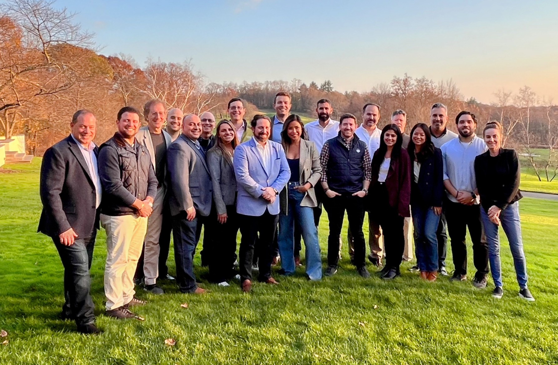Group of professionals posing outdoors on a sunny day, standing on green grass with trees and a clear sky in the background.